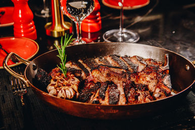Grilled bone-in steak with deep char marks, sliced in a copper pan next to a roasted garlic bulb and rosemary sprig on a dinner table with wine glasses and red plates.