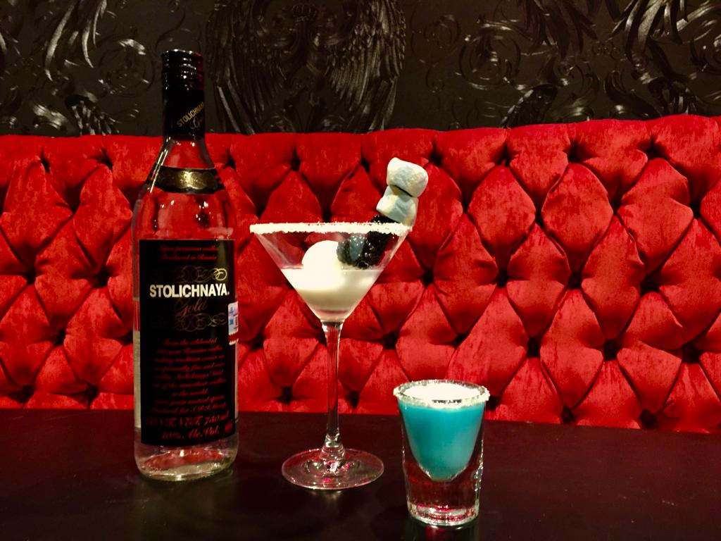 Creamy white martini with marshmallow garnish, a bright blue sugared shot, and a clear vodka bottle on a bar table against a tufted red velvet booth