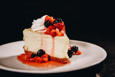 Decadent creamy cheesecake slice on a white plate, drizzled with vibrant mixed-berry compote and syrup and topped with a dollop of whipped cream against a dark background