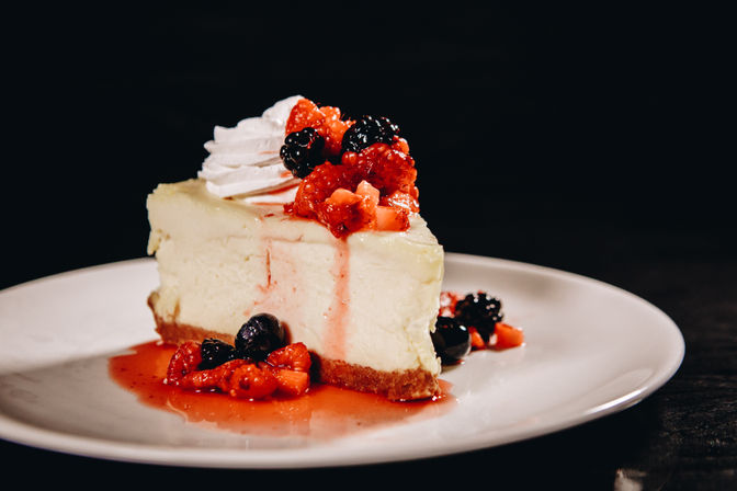 Decadent creamy cheesecake slice on a white plate, drizzled with vibrant mixed-berry compote and syrup and topped with a dollop of whipped cream against a dark background
