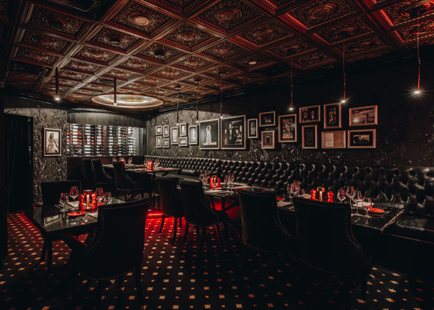 Moody speakeasy-style dining room with tufted black leather banquettes, ornate pressed-tin ceiling, wine bottle wall, framed photos and red-accent candlelit tables set with wine glasses.