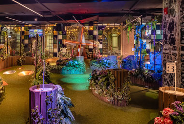 Indoor mini golf course with illuminated windmill, artificial turf pathways, wooden planters, lush faux foliage and colorful mood lighting, family-friendly entertainment