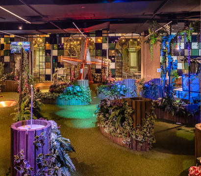 Indoor mini golf course with illuminated windmill, artificial turf pathways, wooden planters, lush faux foliage and colorful mood lighting, family-friendly entertainment