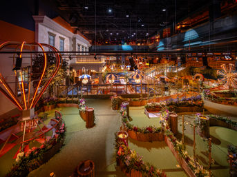 Indoor mini-golf course with winding garden-lined putting greens, neon hot-air-balloon lights, striped umbrella seating, wooden railings and multilevel balconies under warm ambient lighting — family-friendly indoor entertainment venue.