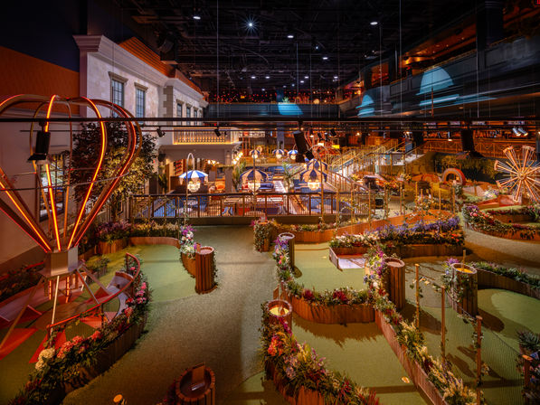 Indoor mini-golf course with winding garden-lined putting greens, neon hot-air-balloon lights, striped umbrella seating, wooden railings and multilevel balconies under warm ambient lighting — family-friendly indoor entertainment venue.