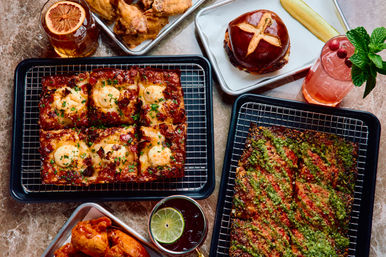 Flat-lay of American comfort food on a marble table: square pan-baked pizza slices with melted cheese and herbs, herb-crusted flatbread, crispy fried chicken wings, a pretzel-bun burger, and two colorful cocktails garnished with citrus and mint.