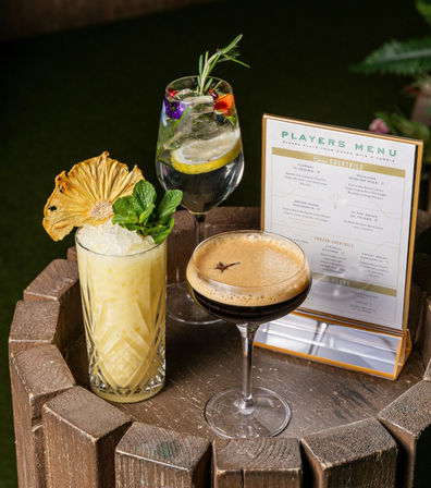 Three craft cocktails on a rustic wooden barrel table in an outdoor patio: a tropical yellow drink with crushed ice, dried pineapple and mint; a flower‑garnished gin & tonic with lemon and rosemary; and a frothy espresso martini beside a tabletop cocktail menu.