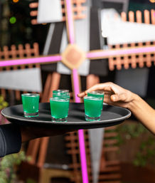 Black serving tray holding six bright green shot glasses, a hand reaching to grab one in a neon-lit bar scene