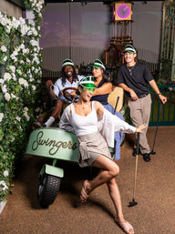 Group of four people wearing green visors posing playfully with a mint-green vintage golf cart and putters at an indoor mini-golf course, white rose wall and decorative clock in the background.