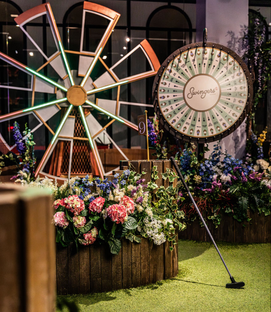 Indoor mini-golf putting green featuring a decorative windmill, round prize wheel, wooden flower planters with pink and purple blooms, and a putter leaning against the planter