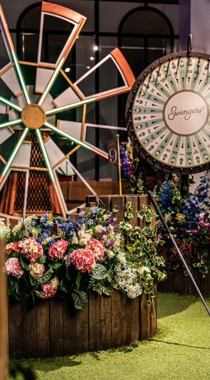 Indoor mini-golf putting green featuring a decorative windmill, round prize wheel, wooden flower planters with pink and purple blooms, and a putter leaning against the planter
