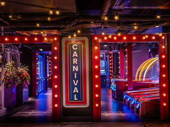 Indoor carnival arcade entrance with vertical marquee reading 'CARNIVAL', warm string bulbs, colorful neon accents and skee-ball lanes