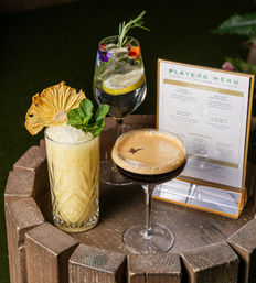 Three cocktails on a rustic wooden barrel: a crushed-ice tropical pineapple tiki garnished with dried pineapple and mint, a floral gin-and-tonic with lemon slice, rosemary and edible flowers, and a creamy espresso martini in a coupe beside a small players menu — outdoor bar summer cocktail scene.