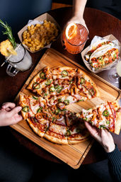 Hand grabbing a jalapeño and red onion pizza slice on a wooden board, surrounded by mac and cheese, soft tacos and colorful cocktails on a dark wood table
