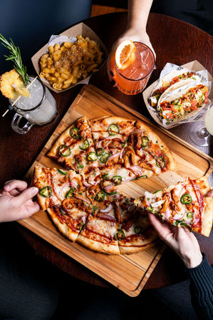 Hand grabbing a jalapeño and red onion pizza slice on a wooden board, surrounded by mac and cheese, soft tacos and colorful cocktails on a dark wood table