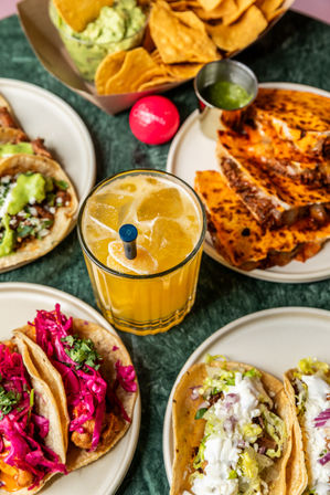 Overhead shot of a Mexican-style spread on a green marble table: orange ice-filled cocktail, tortilla chips with guacamole, grilled quesadilla slices, and assorted street-style tacos topped with pickled red cabbage, shredded lettuce, and crema.