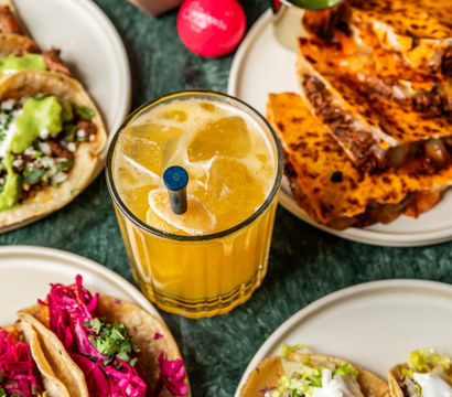 Overhead shot of a Mexican-style spread on a green marble table: orange ice-filled cocktail, tortilla chips with guacamole, grilled quesadilla slices, and assorted street-style tacos topped with pickled red cabbage, shredded lettuce, and crema.