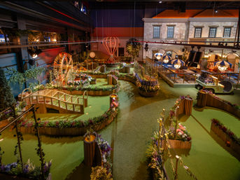 Whimsical indoor mini golf course with green turf, wooden bridges, flower-filled planters, Ferris-wheel and hot-air balloon sculptures, and a covered seating area — family entertainment venue.