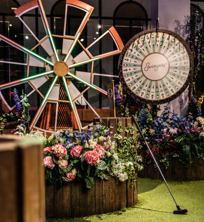 Indoor mini-golf scene with a decorative windmill and large spinning prize wheel, colorful flower-filled wooden planters, a putter resting on artificial turf and a small flag marked 9.