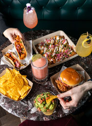 Overhead shot of a marble restaurant table with tacos, nacho chips and guacamole, loaded fries topped with pickled onions and crema, a fried chicken sandwich and colorful cocktails, two hands reaching in.