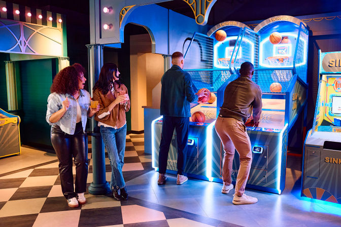 Four friends in a neon-lit indoor arcade: two men shooting hoops at electronic basketball skee-ball machines while two women cheer with drinks on a checkered floor.