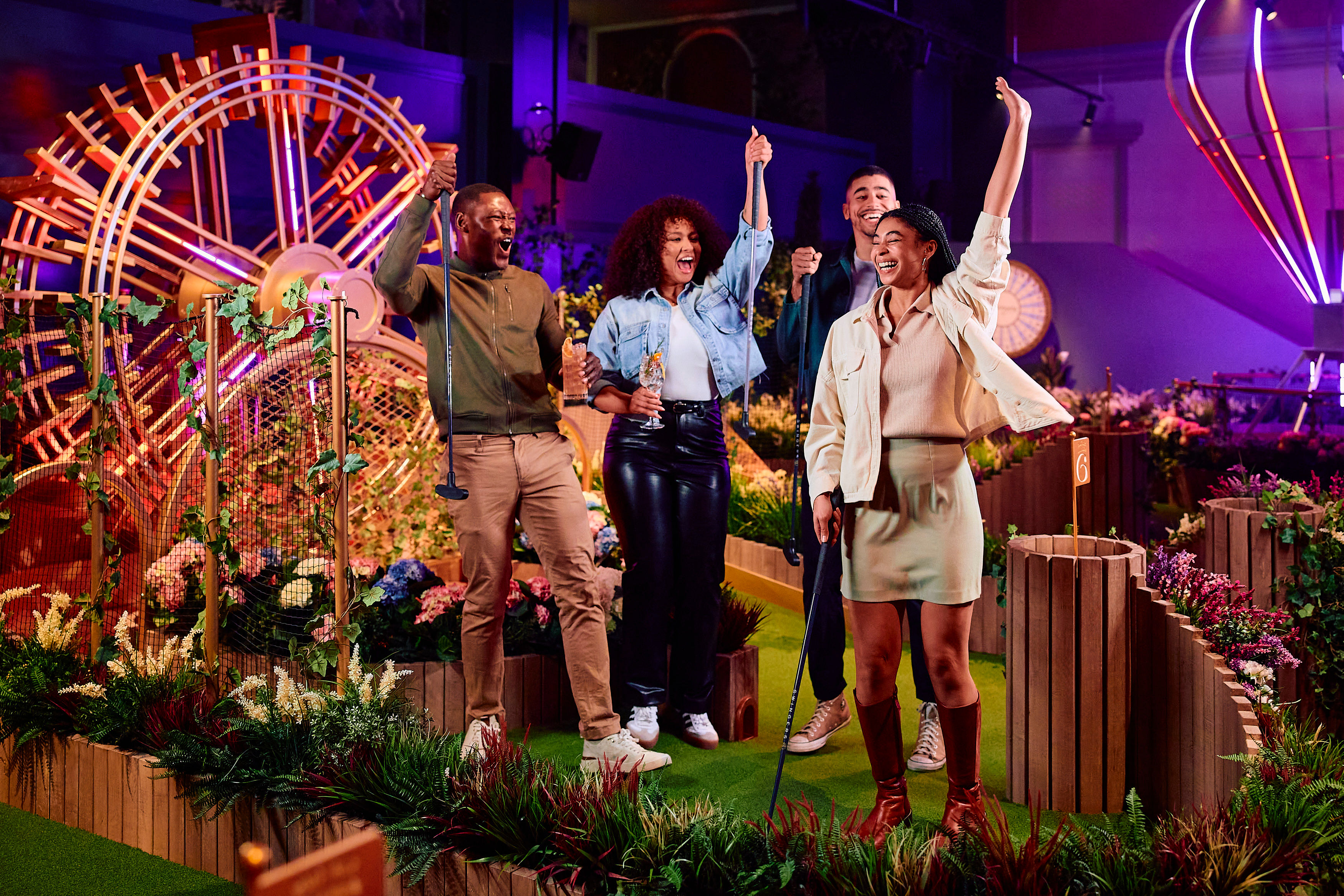 Group of four adults cheering on a neon-lit indoor mini golf course, raising putters and drinks amid planter boxes, colorful flowers, and glowing decorative art.