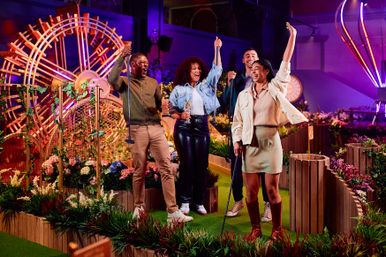 Group of four adults cheering on a neon-lit indoor mini golf course, raising putters and drinks amid planter boxes, colorful flowers, and glowing decorative art.