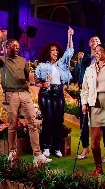 Group of four adults cheering on a neon-lit indoor mini golf course, raising putters and drinks amid planter boxes, colorful flowers, and glowing decorative art.