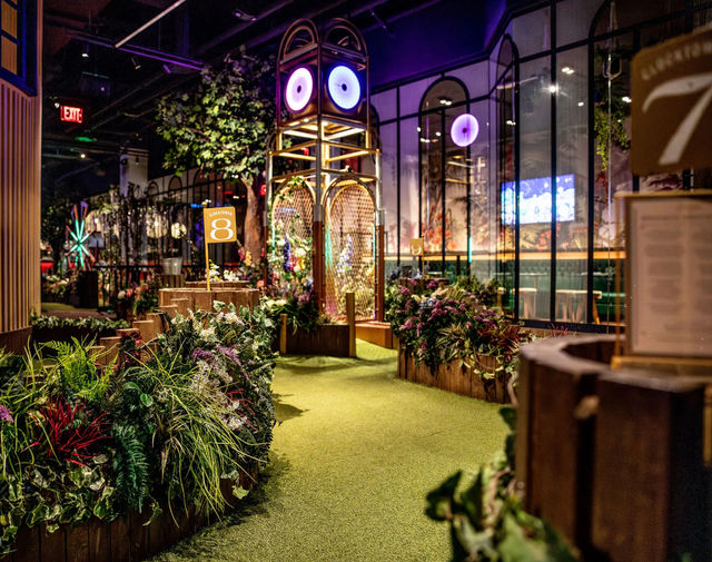 Indoor glow-lit mini golf hole with artificial turf, lush tropical planters, wooden course sign and a whimsical tower with purple neon 'eyes' in an urban entertainment venue