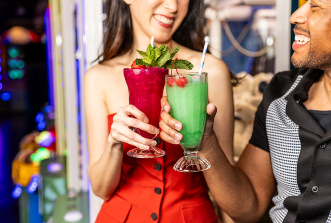 Two people clinking colorful cocktails at a lively bar — magenta mint-garnished frozen drink and green cherry-topped blended cocktail, smiling