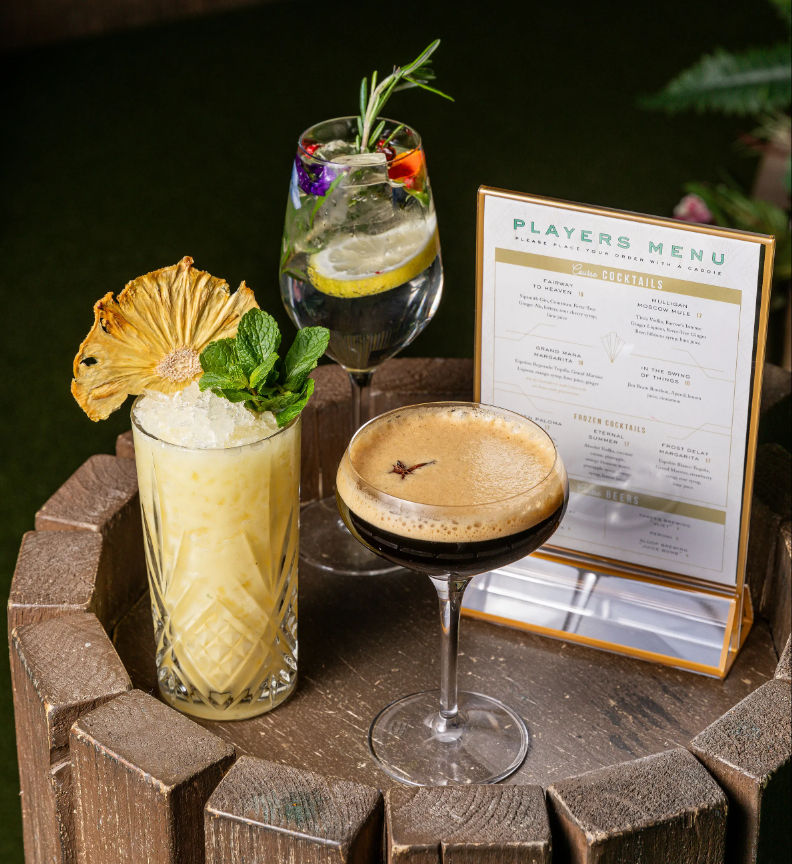 Three colorful cocktails arranged on a round wooden barrel table outdoors with a printed menu: a tiki-style pineapple drink with crushed ice, mint and dried pineapple garnish; a floral gin-and-tonic with lemon and rosemary; and a creamy espresso martini topped with foam and star anise.