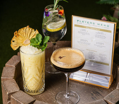 Three colorful cocktails arranged on a round wooden barrel table outdoors with a printed menu: a tiki-style pineapple drink with crushed ice, mint and dried pineapple garnish; a floral gin-and-tonic with lemon and rosemary; and a creamy espresso martini topped with foam and star anise.
