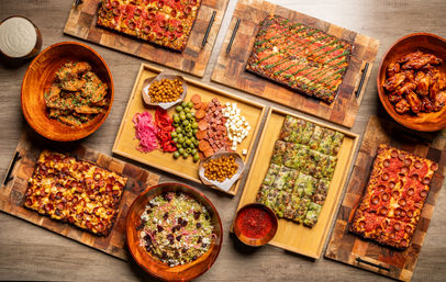 Overhead party spread on rustic wooden boards: rectangular pan pizzas, saucy chicken wings, a mixed salad, and a charcuterie-style tray with olives, cheeses, pepperoni, pickled veggies, chickpea snacks and marinara dip.