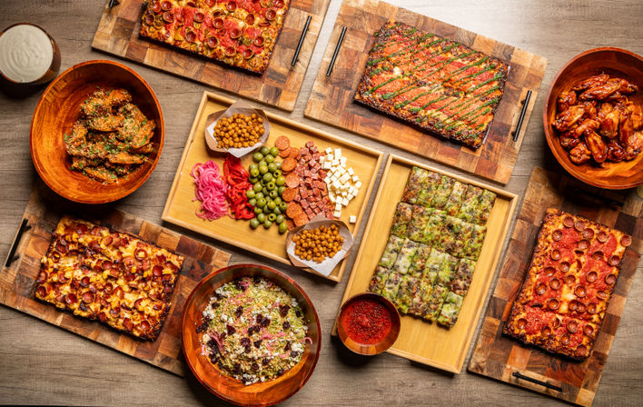 Overhead party spread on rustic wooden boards: rectangular pan pizzas, saucy chicken wings, a mixed salad, and a charcuterie-style tray with olives, cheeses, pepperoni, pickled veggies, chickpea snacks and marinara dip.