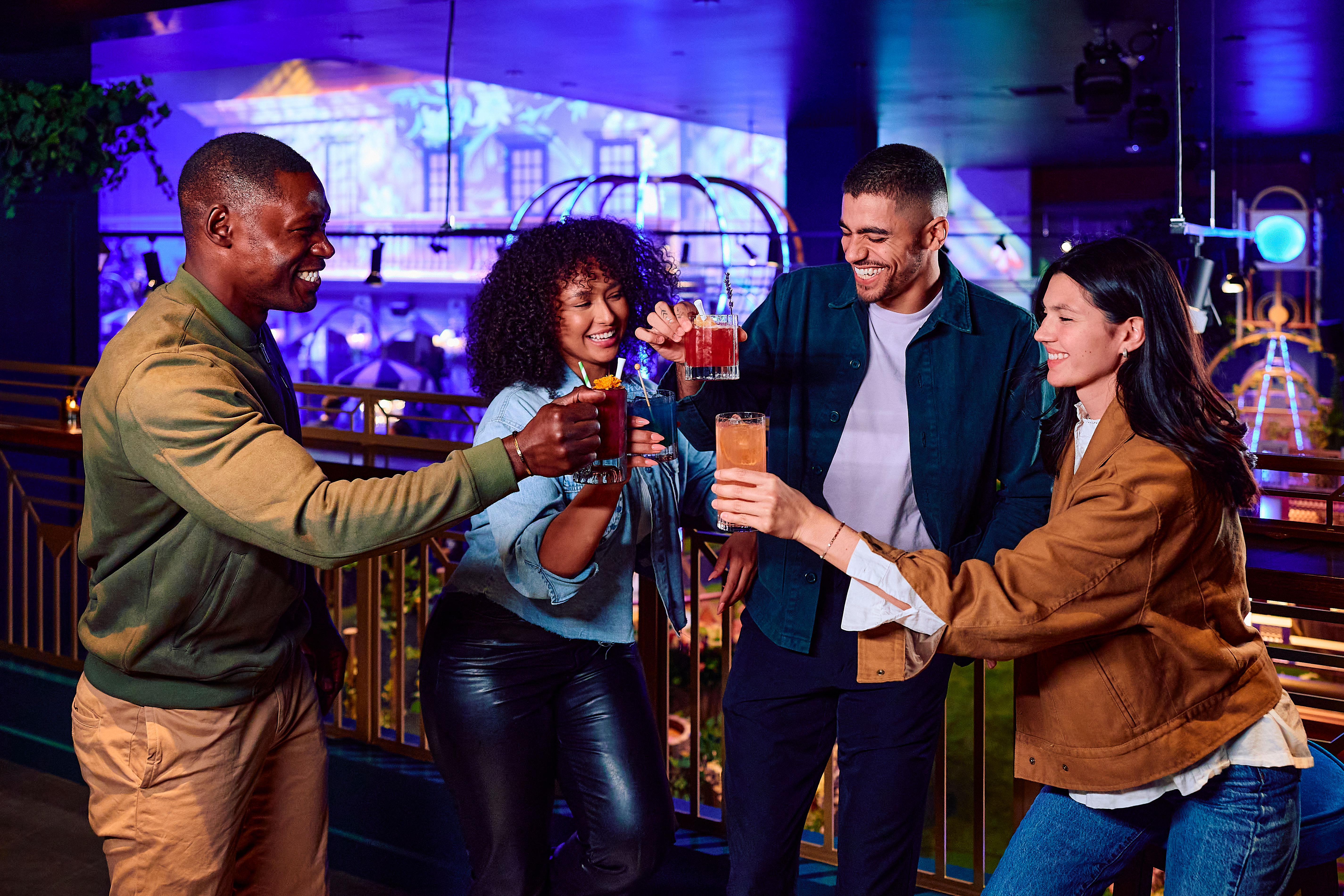 Four friends laughing and toasting colorful cocktails on a neon-lit indoor bar balcony, enjoying lively urban nightlife