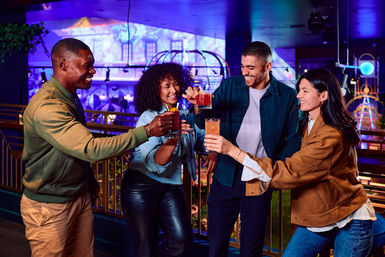 Four friends laughing and toasting colorful cocktails on a neon-lit indoor bar balcony, enjoying lively urban nightlife