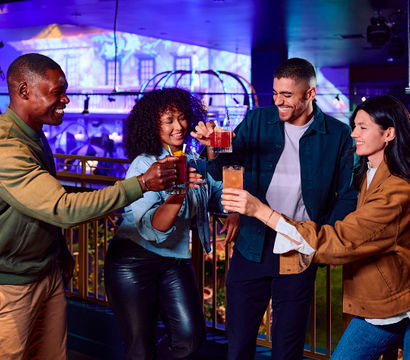 Four friends laughing and toasting colorful cocktails on a neon-lit indoor bar balcony, enjoying lively urban nightlife