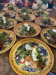 Vibrant Talavera-style plates on a counter, each holding a Mexican sope topped with shredded meat, shredded lettuce, crumbled cheese and dollops of crema, with onions and fruit in the background.
