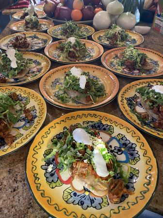 Vibrant Talavera-style plates on a counter, each holding a Mexican sope topped with shredded meat, shredded lettuce, crumbled cheese and dollops of crema, with onions and fruit in the background.