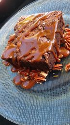 Close-up of a fudgy chocolate brownie drizzled with caramel and scattered pecans on a textured blue plate, decadent dessert shot.