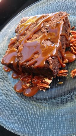 Close-up of a fudgy chocolate brownie drizzled with caramel and scattered pecans on a textured blue plate, decadent dessert shot.