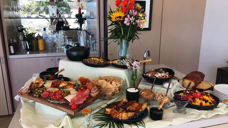 Indoor brunch buffet on a kitchen island featuring a charcuterie board with cheeses, cured meats, croissants and olives, bowls of tortilla chips, granola and nuts, French toast and sliced loaf bread, a colorful fresh fruit salad, assorted pastries and a tall vase of sunflowers and mixed flowers.