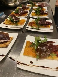 Rows of party-ready appetizers on white square plates: rectangular flatbread topped with roasted squash, peppery arugula, thin slices of cured meat and balsamic glaze on a stainless-steel kitchen counter.
