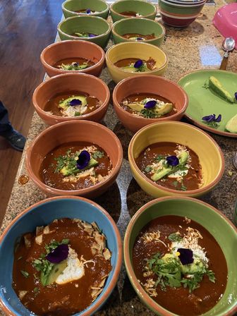 Rows of colorful ceramic bowls on a kitchen counter filled with rich Mexican-style mole topped with sliced avocado, crumbled cheese, cilantro and purple edible flowers
