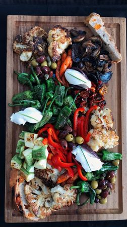 Overhead shot of a colorful Mediterranean-style roasted vegetable board on a wooden board with charred cauliflower, blistered padrón peppers, grilled zucchini, sautéed mushrooms, roasted red peppers, mixed olives, dollops of creamy burrata-style cheese, roasted carrots and toasted bread — shareable appetizer platter.