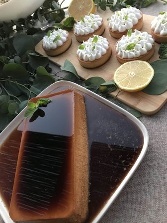 Glossy caramel flan in a white dish garnished with mint, beside mini lemon tarts topped with piped meringue and mint on a wooden board, fresh lemon halves and eucalyptus on a rustic linen table — summer dessert spread.