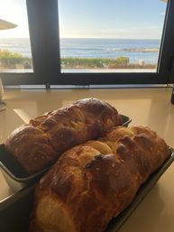 Sunlit coastal kitchen scene with two golden braided loaves cooling in loaf pans on a counter and a large window framing ocean waves and a seaside patio.