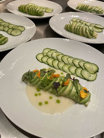 Fine-dining plates of thinly sliced cucumber ribbons and an avocado-wrapped roll topped with microgreens, yellow edible flowers and green oil dots, arranged on white plates in a professional kitchen.