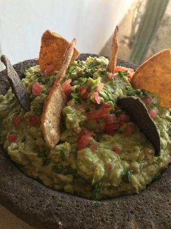 Creamy molcajete guacamole in a volcanic stone bowl topped with pico de gallo and sliced scallions, served with blue and yellow tortilla chips