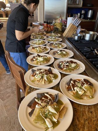 Multiple plates of chilaquiles with green and red salsa, crema and crumbled cheese on blue and yellow tortilla chips lined up on a granite kitchen island while a cook finishes plating — Mexican breakfast/brunch prep.